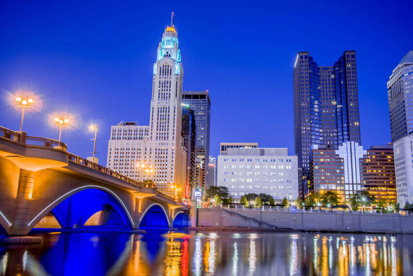 Cityscape of a downtown area with illuminated buildings, a bridge over a reflective river, and a clear evening sky.