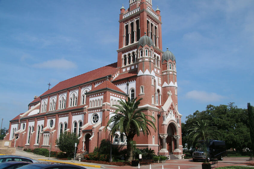 A red-brick church with a tall, ornate tower and arched windows, surrounded by palm trees and parked cars on a sunny day.