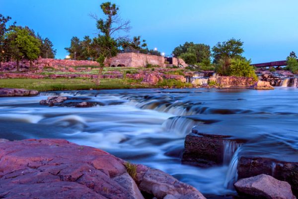 A serene river flows over multiple small waterfalls, surrounded by green trees and rocky terrain under a clear sky during twilight.