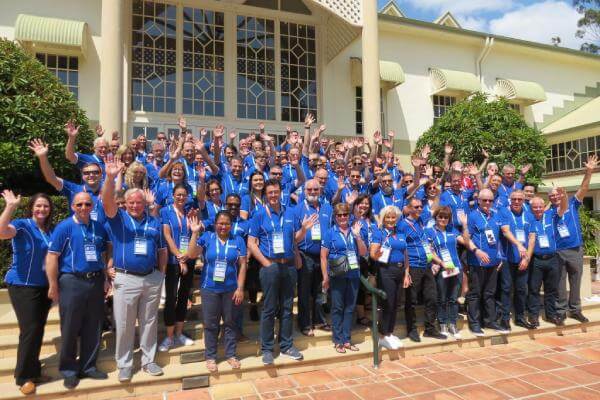 A large group of people wearing blue shirts and name tags stand on the steps of a building, smiling and waving at the camera. A large group of people wearing blue shirts stand on the steps of a building, smiling and waving at the camera.