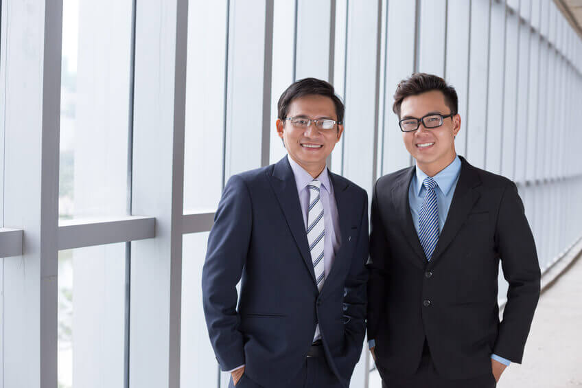 Two men in matching dark suits stand side by side in a modern office corridor with large windows. They are both smiling and looking at the camera. Two men in matching dark suits stand side by side in a modern office corridor with large windows. They are both smiling and looking at the camera.