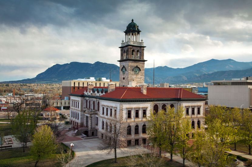 A historic building with a clock tower sits in front of a mountainous landscape under a cloudy sky. Trees surround the building.