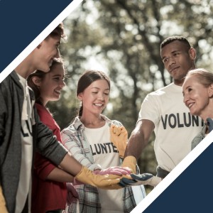 A diverse group of people wearing volunteer shirts stand in a circle outdoors, placing their hands together in a show of unity.