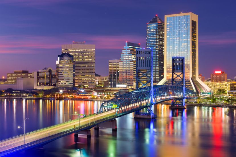 City skyline at dusk with illuminated skyscrapers and a lit-up bridge reflecting on the water.