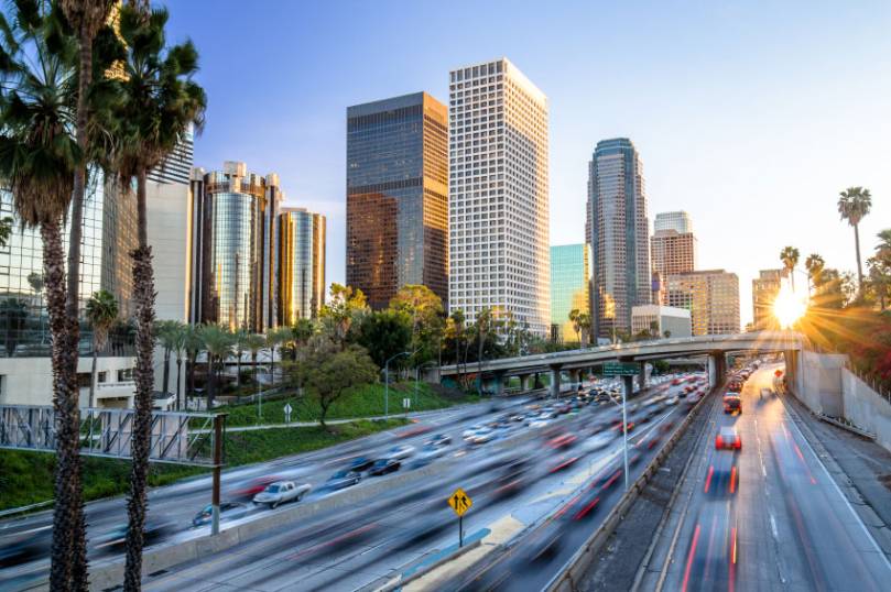 Sunset view of a busy freeway with blurred traffic in front of downtown skyscrapers and palm trees in Los Angeles.