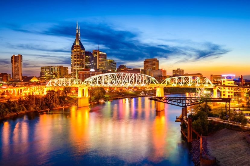 A city skyline at sunset with a prominent bridge over a river, reflecting colorful lights and skyscrapers in the background.