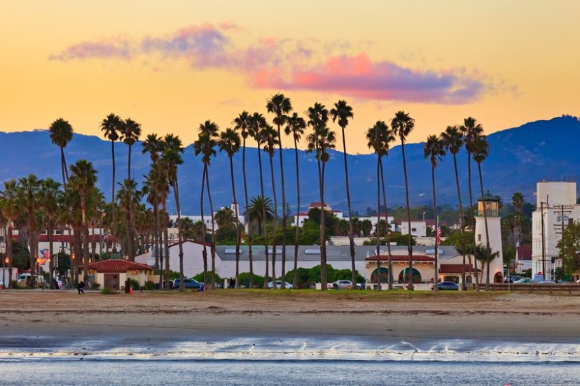 A beach scene at sunset with tall palm trees, buildings in the background, and mountains under a colorful sky.
