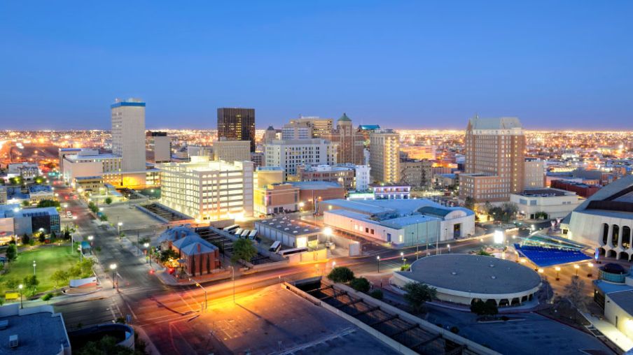 Aerial view of a cityscape at dusk, featuring illuminated buildings, streets, and a visible horizon.
